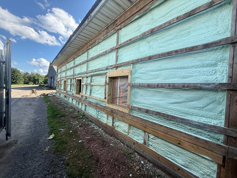 Large agricultural building with turquoise-painted metal siding and wooden frame under construction or renovation, set against blue sky